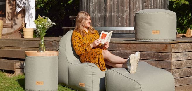 eine Frau die auf grünen Polstern in ihrem Garten ein Buch liest.